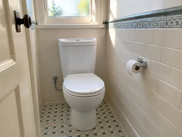 Clean white toilet in a small tiled bathroom with window and patterned trim in Arncliffe